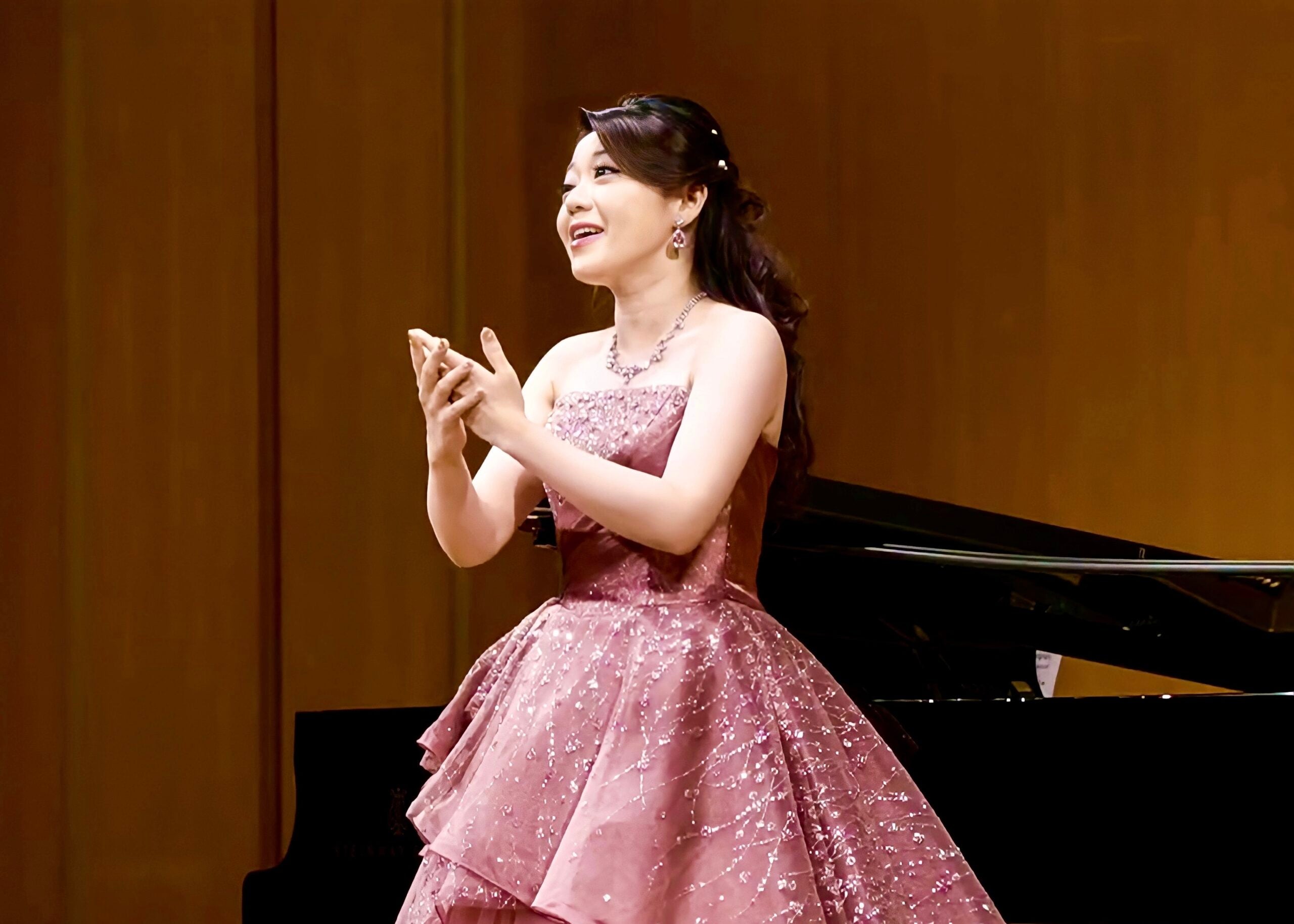 The scientist, wearing a pink gown, sings opera in front of a piano.